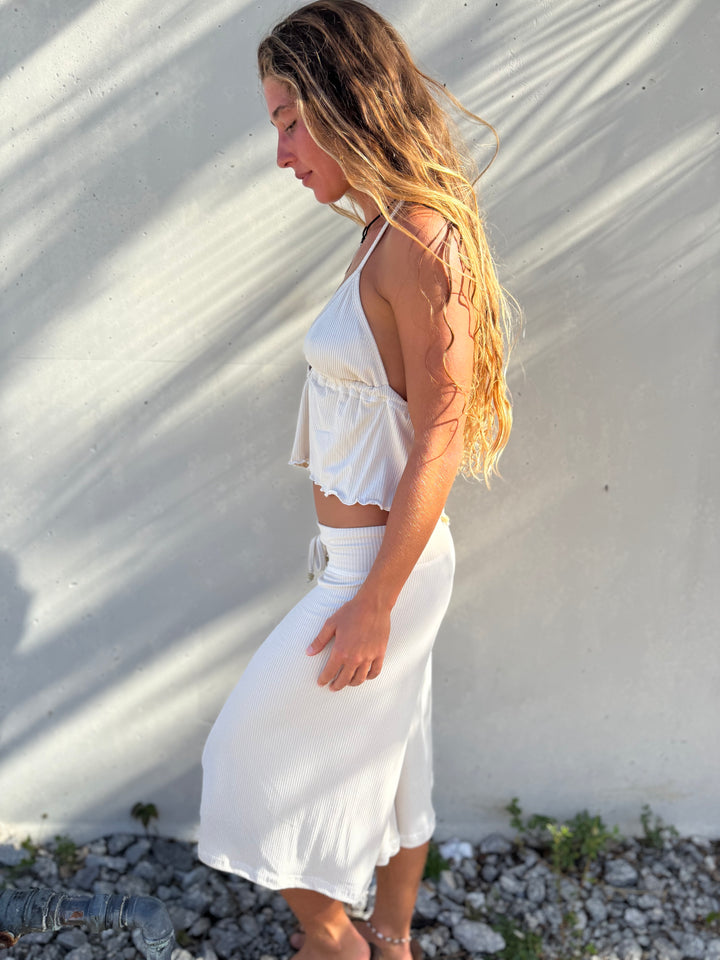 A woman with long blonde hair stands in profile against a sunlit concrete wall, wearing a white sleeveless crop top and a matching white midi skirt.