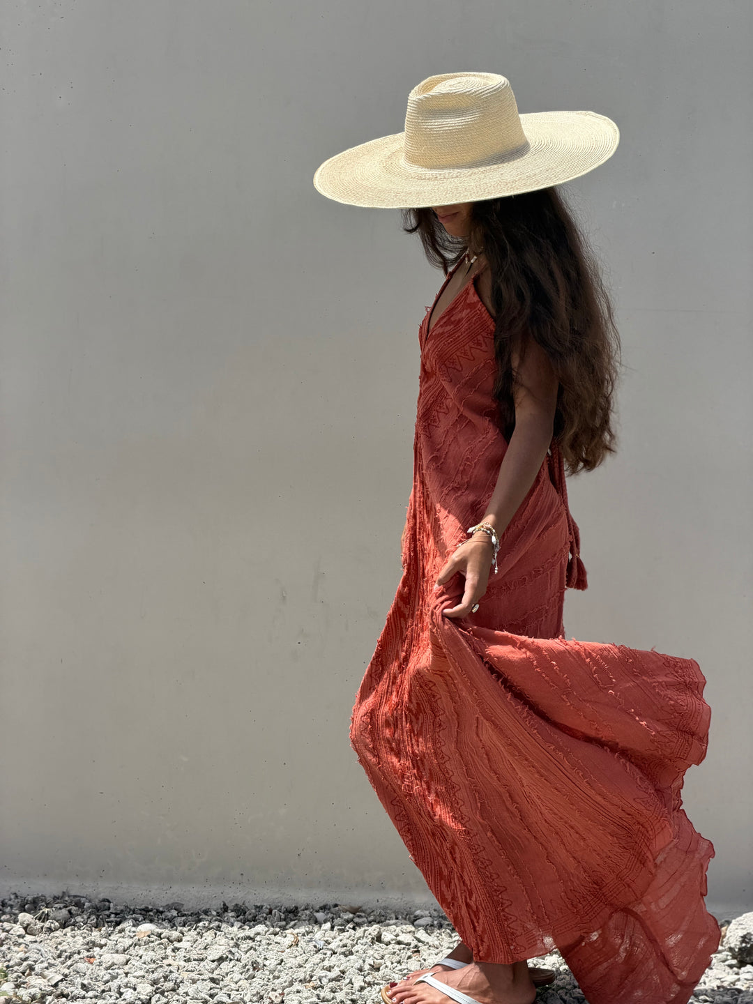 A woman wearing a wide-brimmed straw hat and a flowing terracotta-colored maxi dress walks along a gravel path against a plain light gray wall.