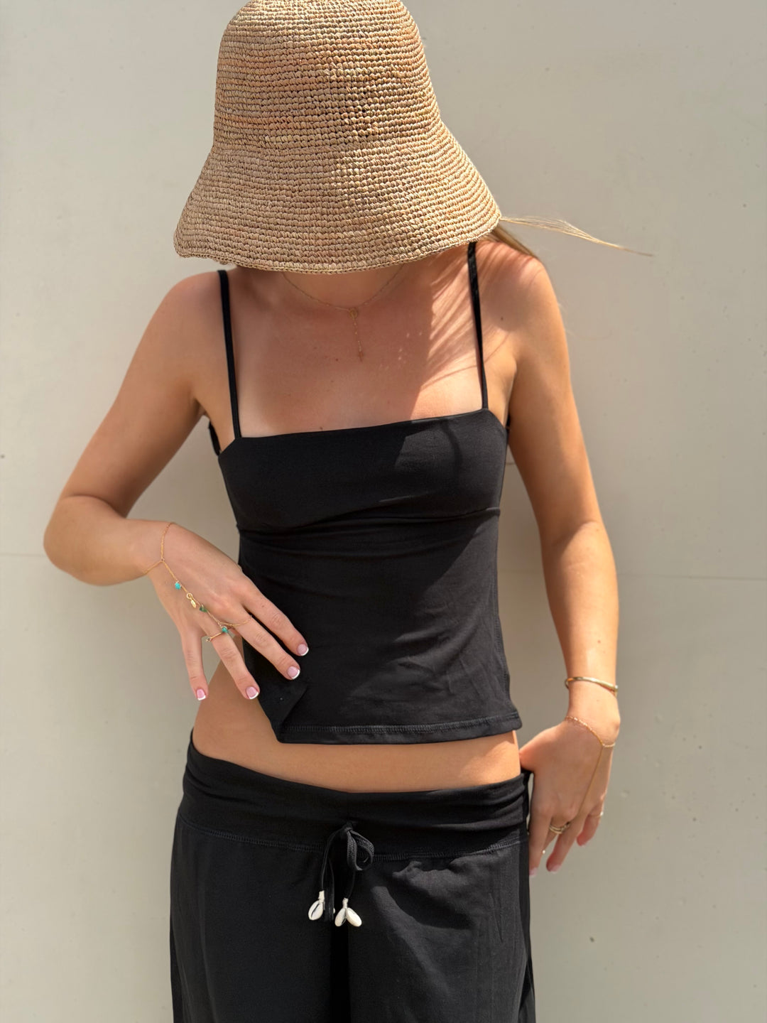 A woman wearing a black spaghetti-strap crop top and black drawstring sweatpants, with a large woven straw bucket hat, standing against a plain light gray wall.