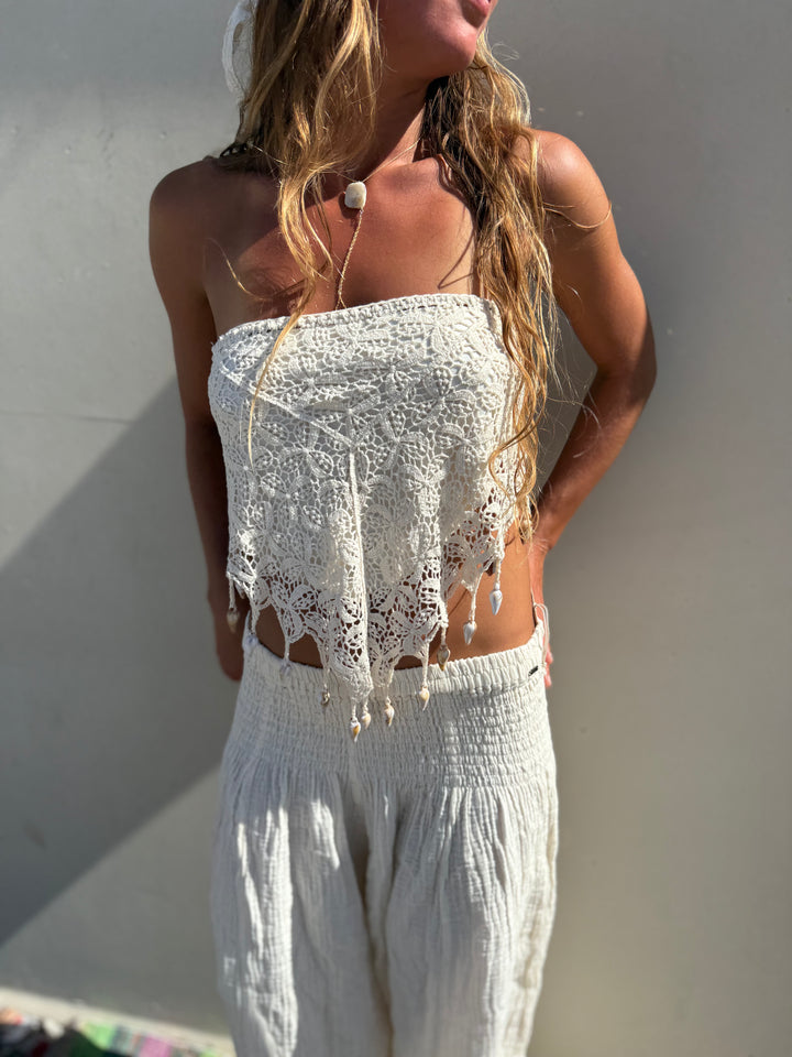 A woman wearing a white crochet strapless tube top with fringe and matching white textured pants, standing against a plain light wall in bright natural light.