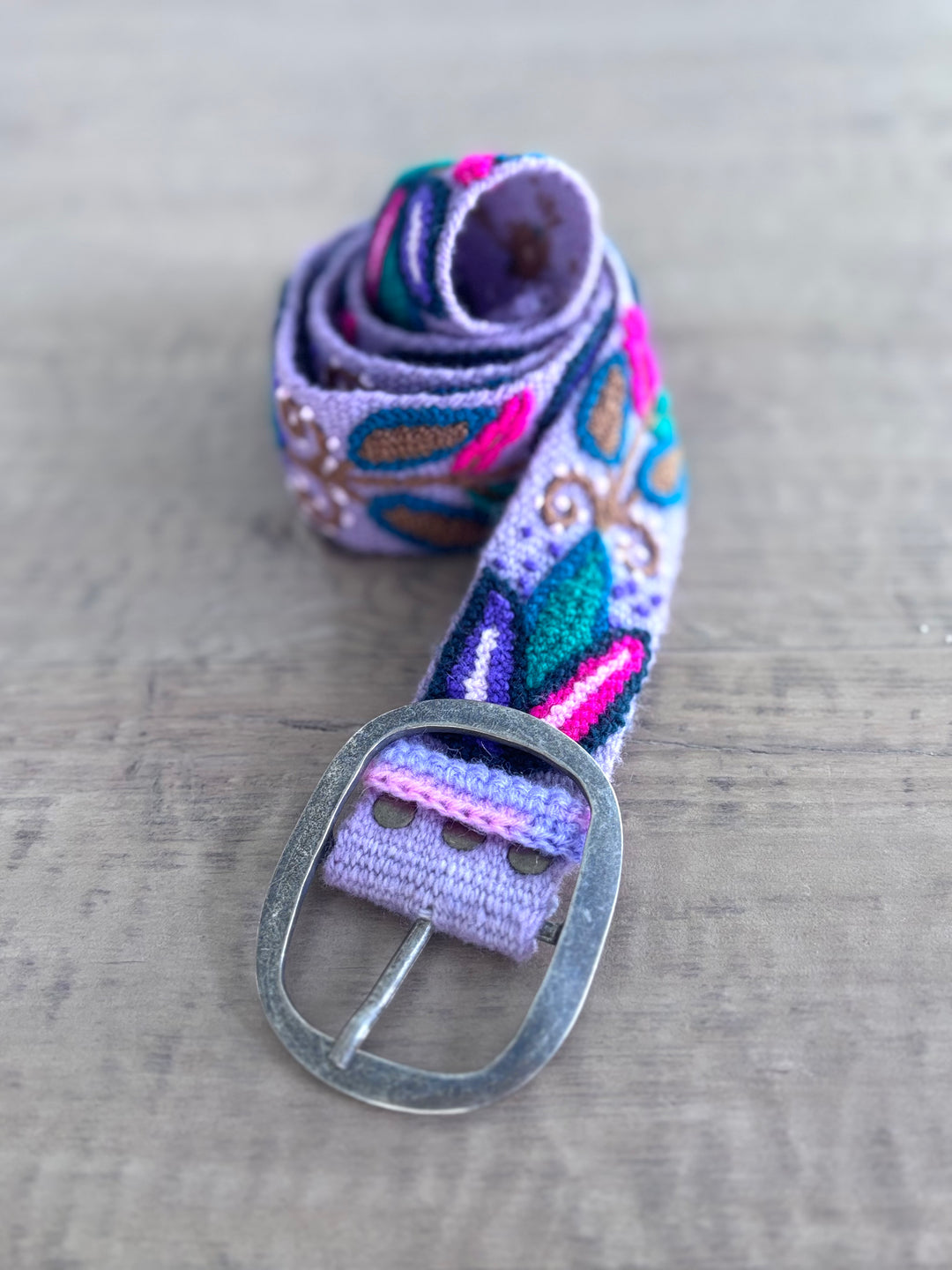 A woven fabric belt with a silver metal buckle, featuring a multicolor pattern of purple, teal, pink, and brown on a light gray wooden background.