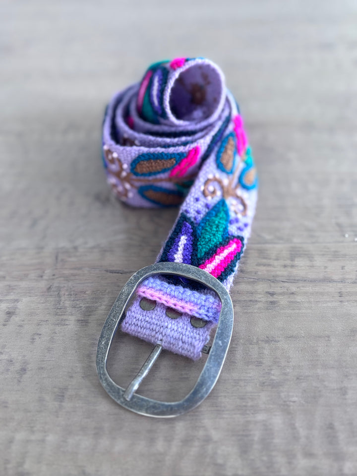 A woven fabric belt with a silver metal buckle, featuring a multicolor pattern of purple, teal, pink, and brown on a light gray wooden background.