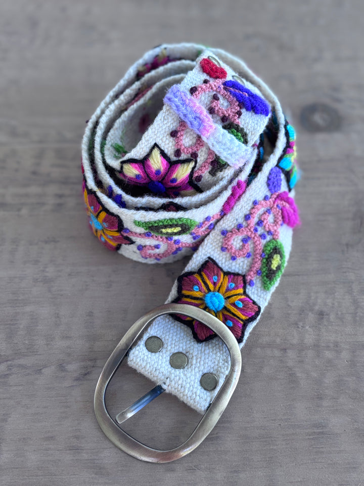 A white woven fabric belt with colorful embroidered floral and geometric patterns, featuring a silver-toned metal buckle, displayed on a gray wooden surface.