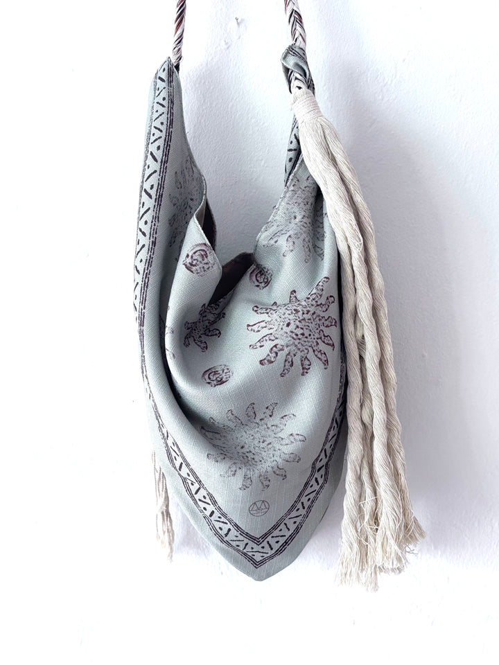 A light gray silk bandana bag with brown sunburst patterns and a geometric border, featuring cream-colored braided shoulder straps with tassels, hanging against a white wall.