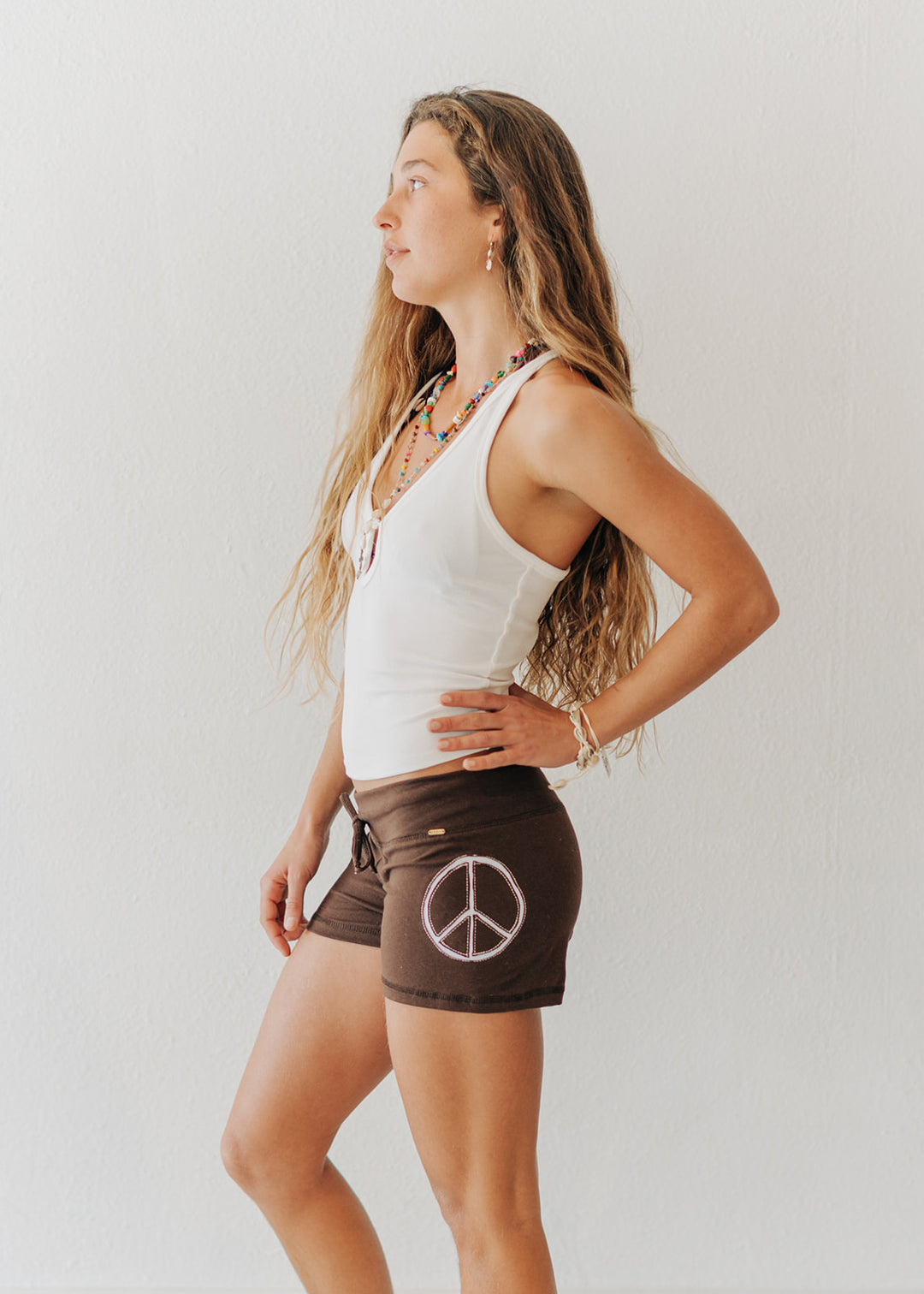 Woman wearing a white tank top and brown shorts with a peace symbol on a plain background