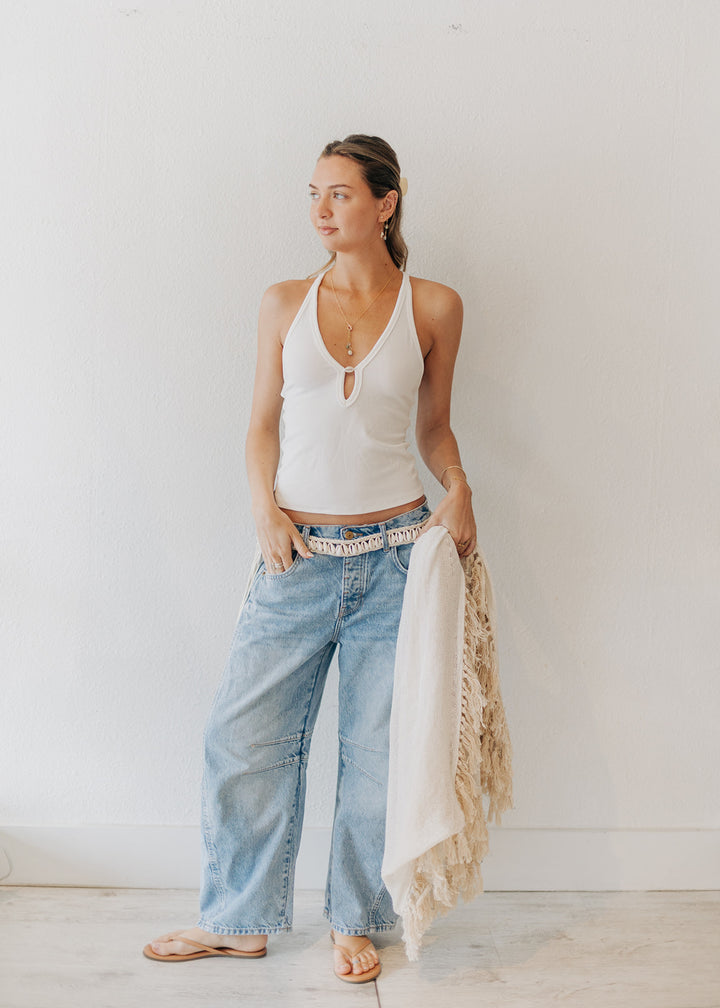Woman wearing a white tank top and blue jeans with a beige scarf, standing against a plain background.