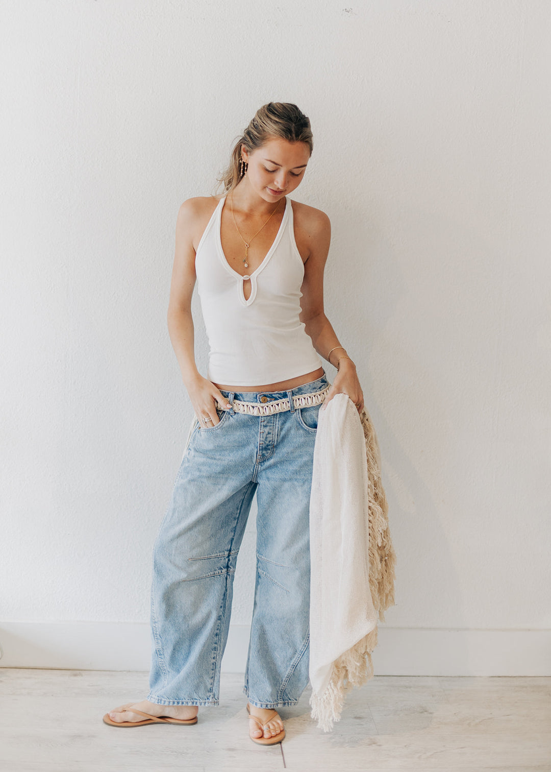 Woman wearing a white tank top and blue jeans with a scarf, standing against a plain background.