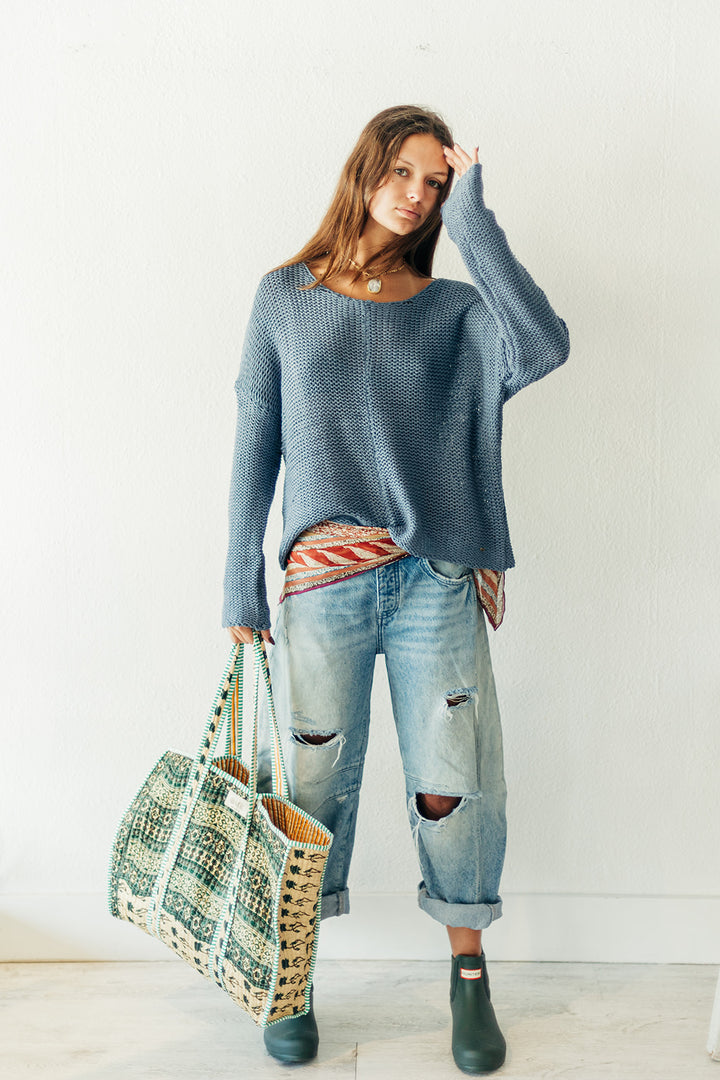 Woman wearing a blue sweater and ripped jeans, holding a patterned bag against a white background