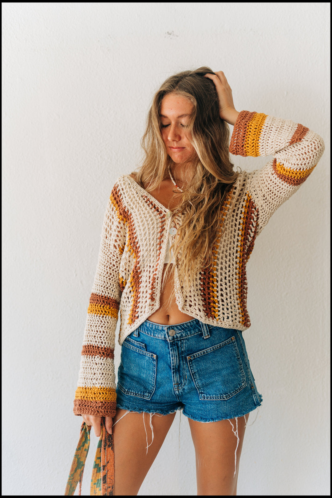 Woman wearing a colorful crochet cardigan and denim shorts against a white background