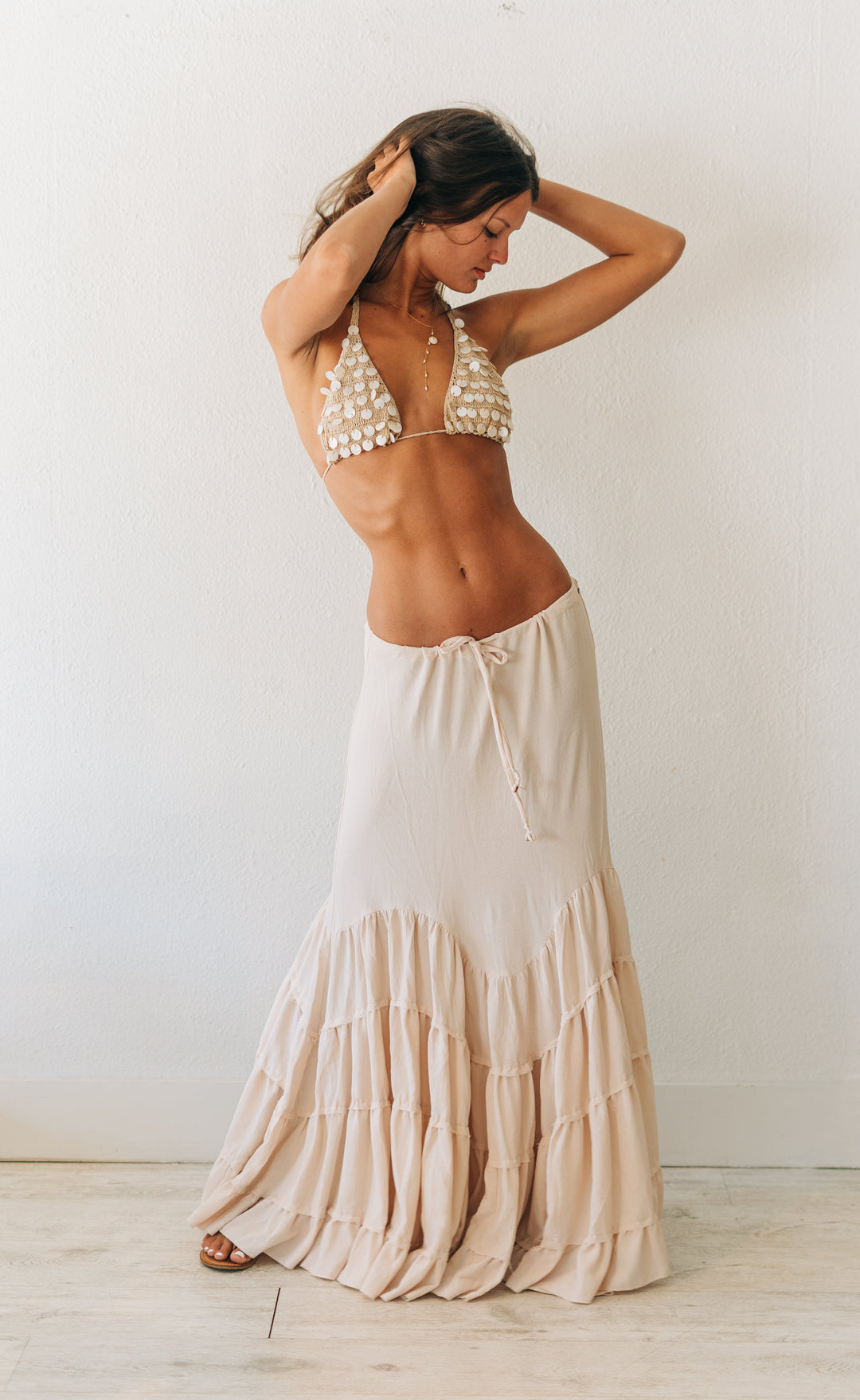 Woman wearing a white bikini top with pearl details and a long beige skirt against a white wall.
