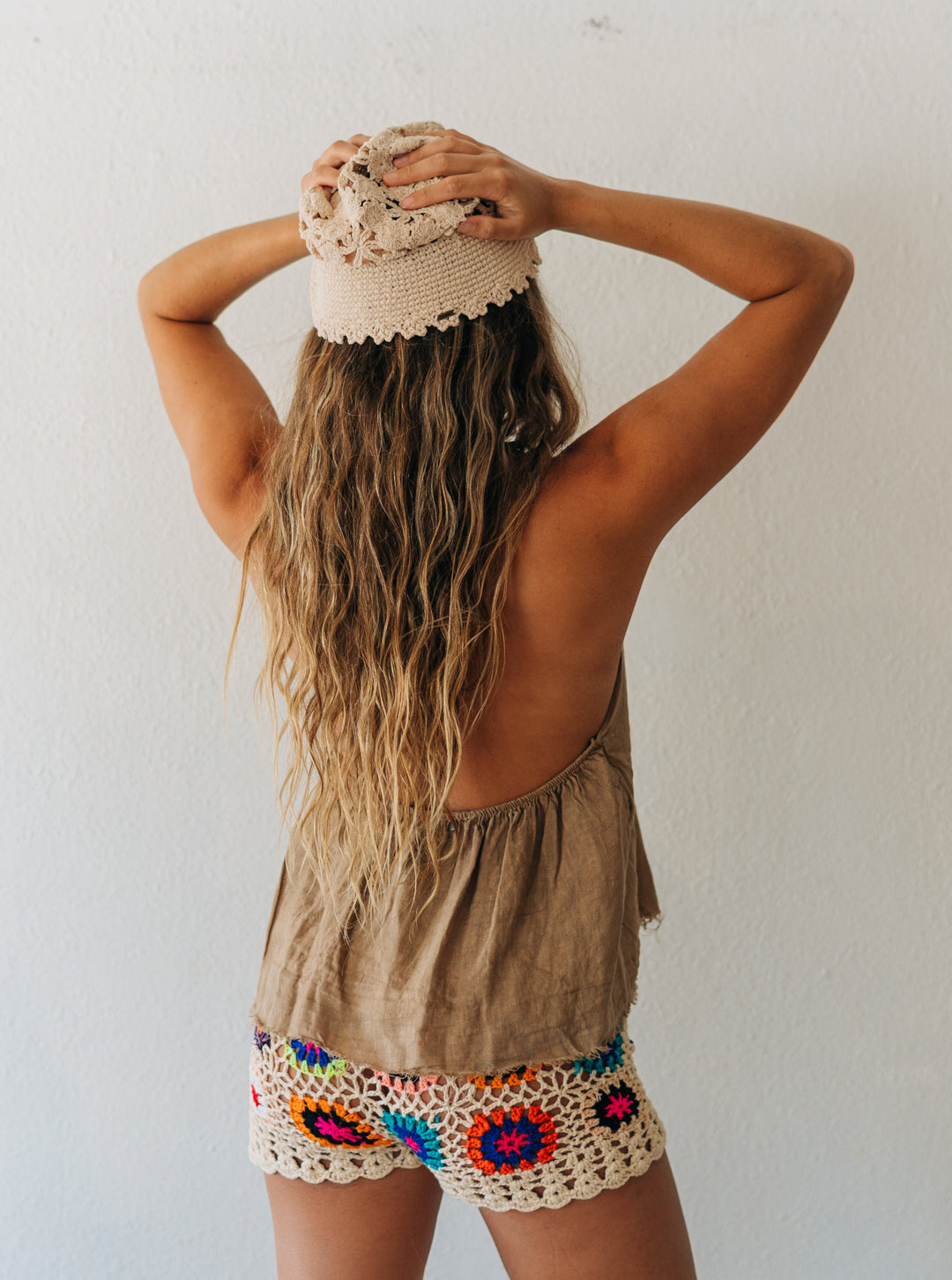 Person wearing a beige sleeveless top and colorful crochet shorts against a white wall.