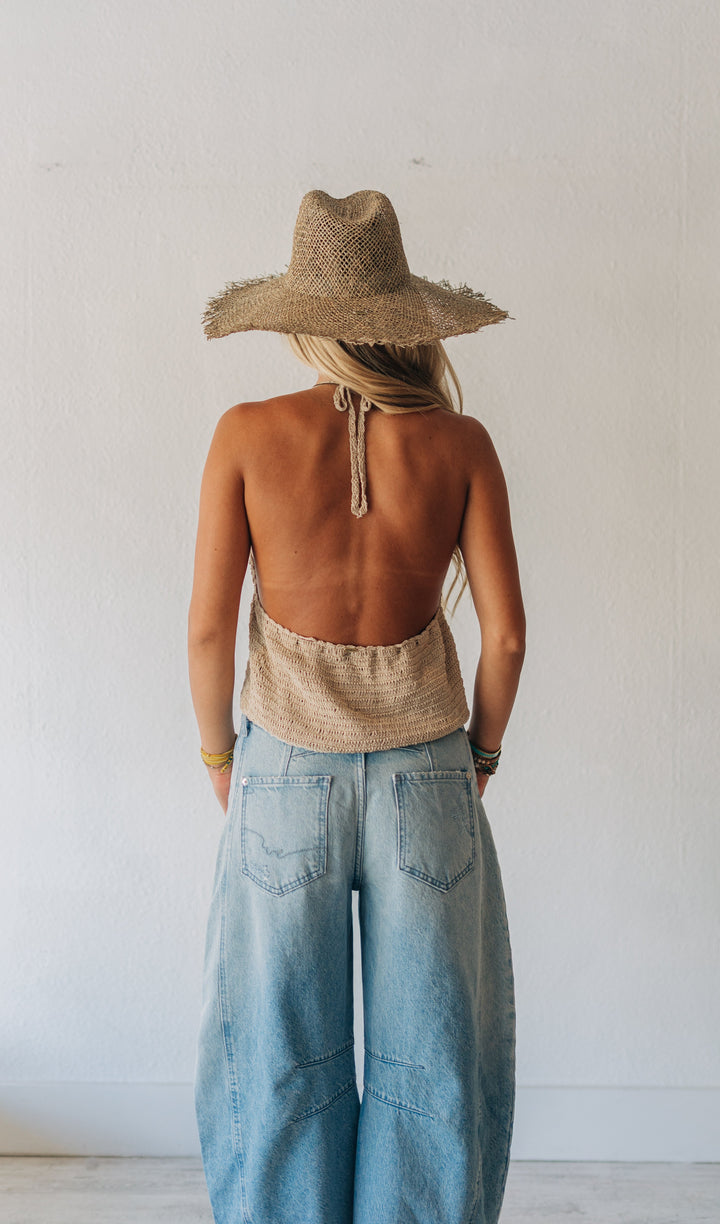 Person wearing a straw hat, sleeveless top, and wide-leg jeans standing against a white wall.