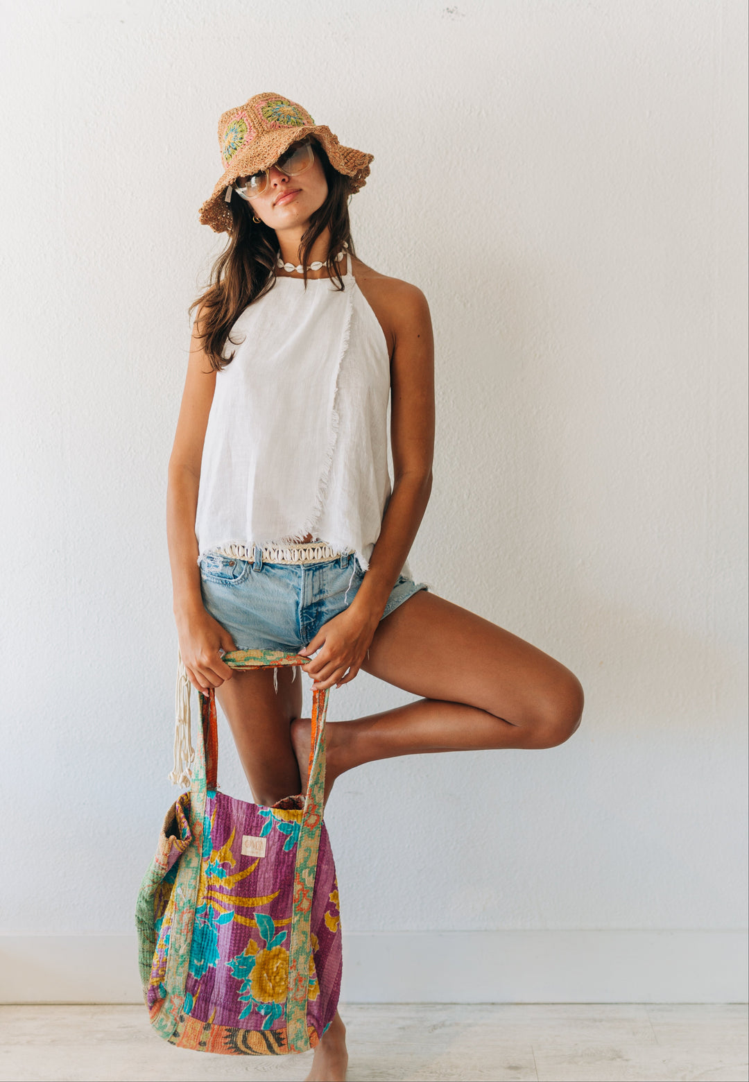 Woman in white halter top and denim shorts holding a colorful bag against a white wall.