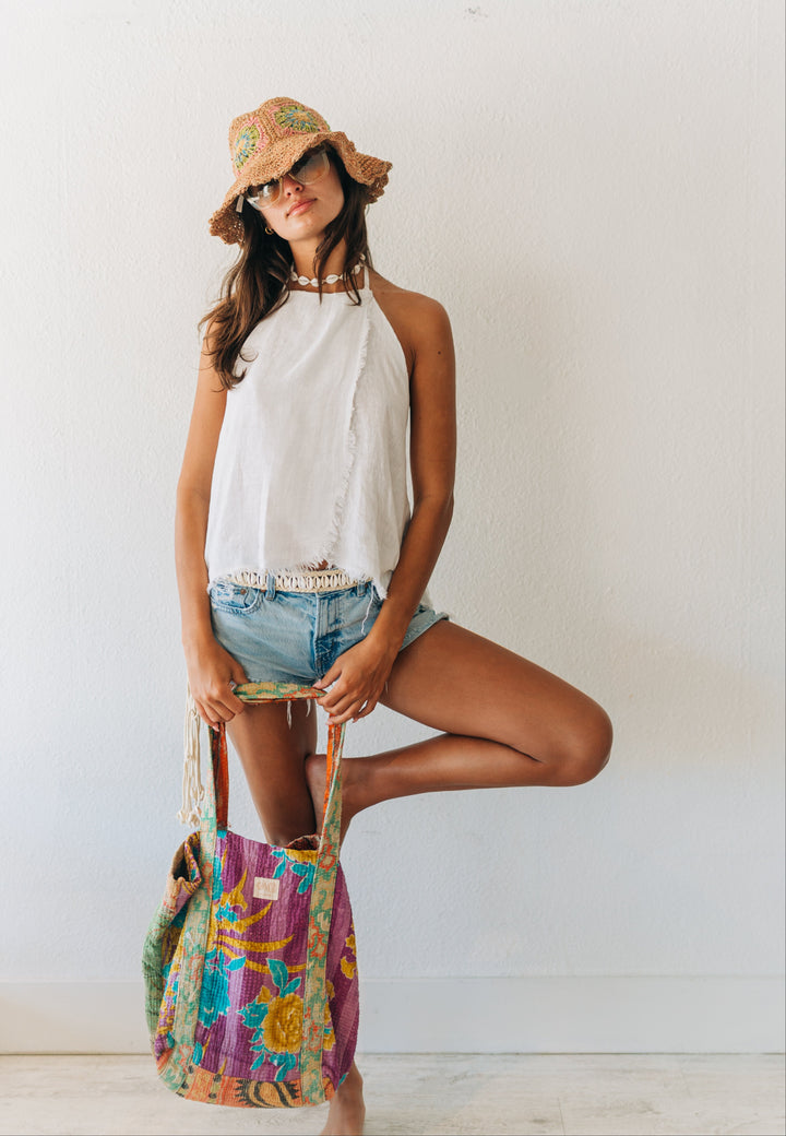 Woman in white halter top and denim shorts holding a colorful bag against a white wall.