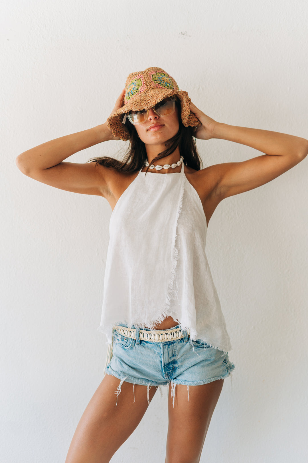 Woman wearing a white halter top, denim shorts, and a straw hat with sunglasses on a plain background