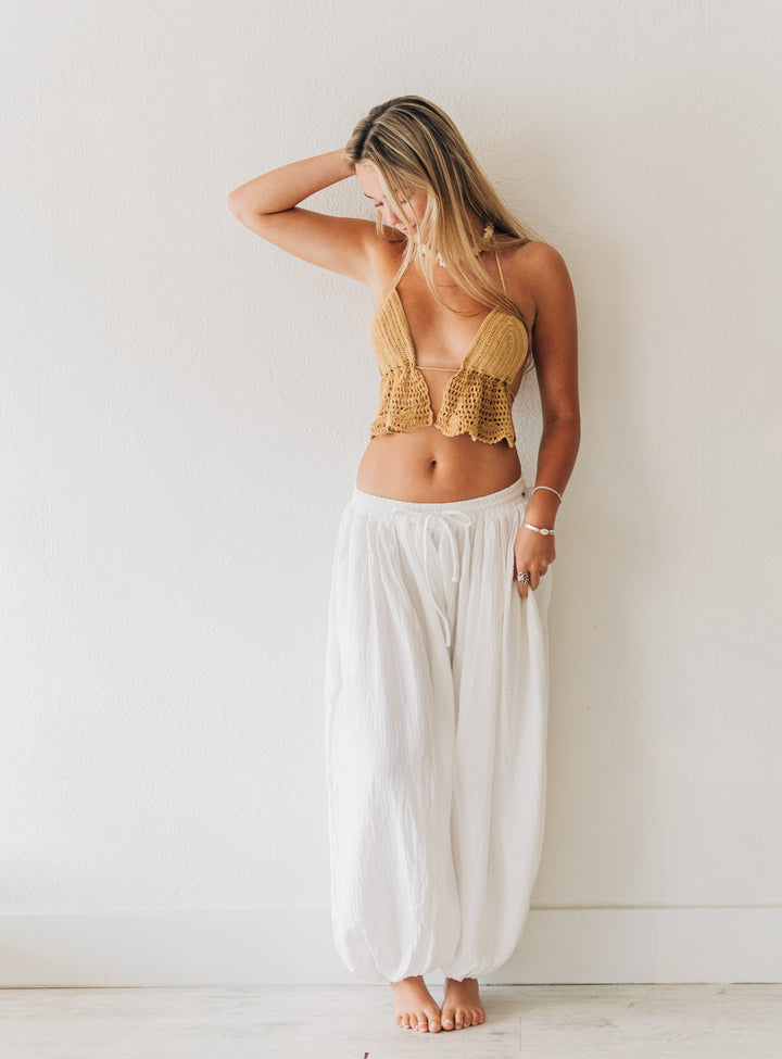 Woman wearing a gold top and white pants standing against a white wall.