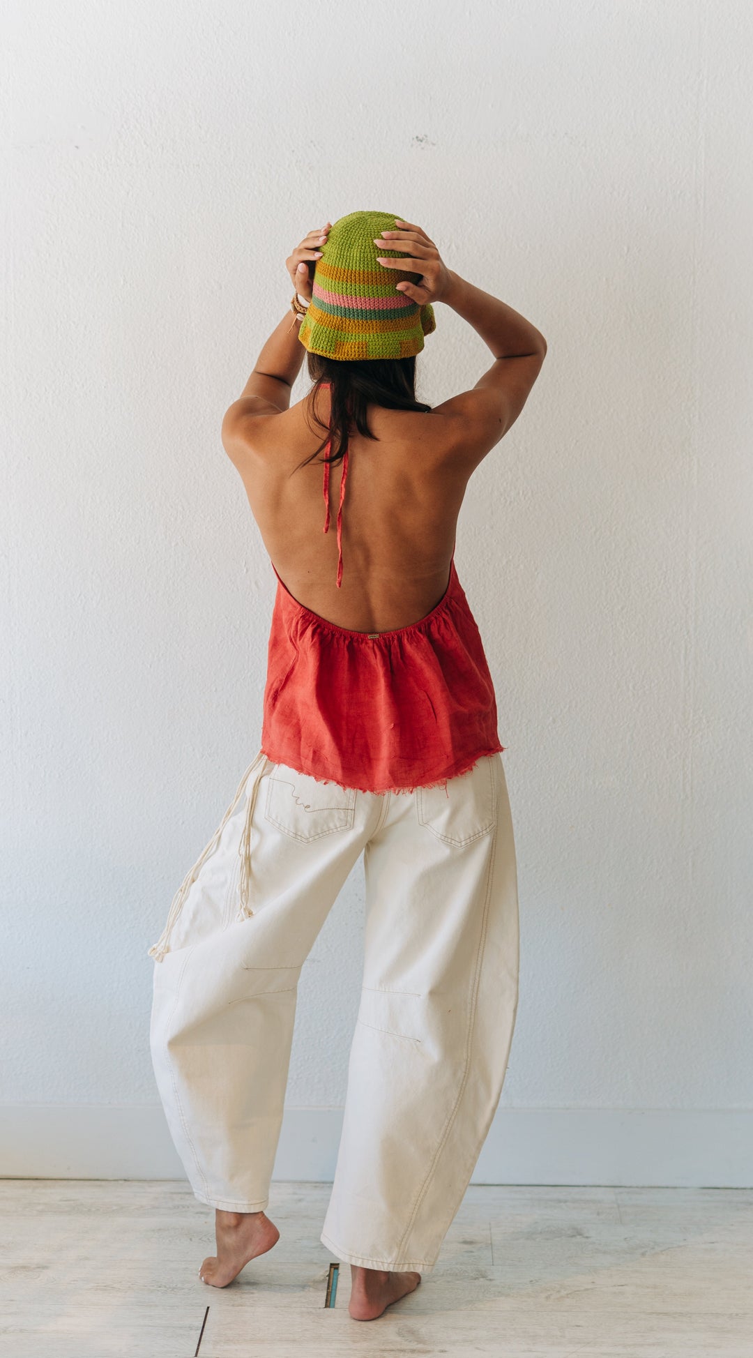 Person wearing a red top and white pants, holding a colorful hat against a white wall.