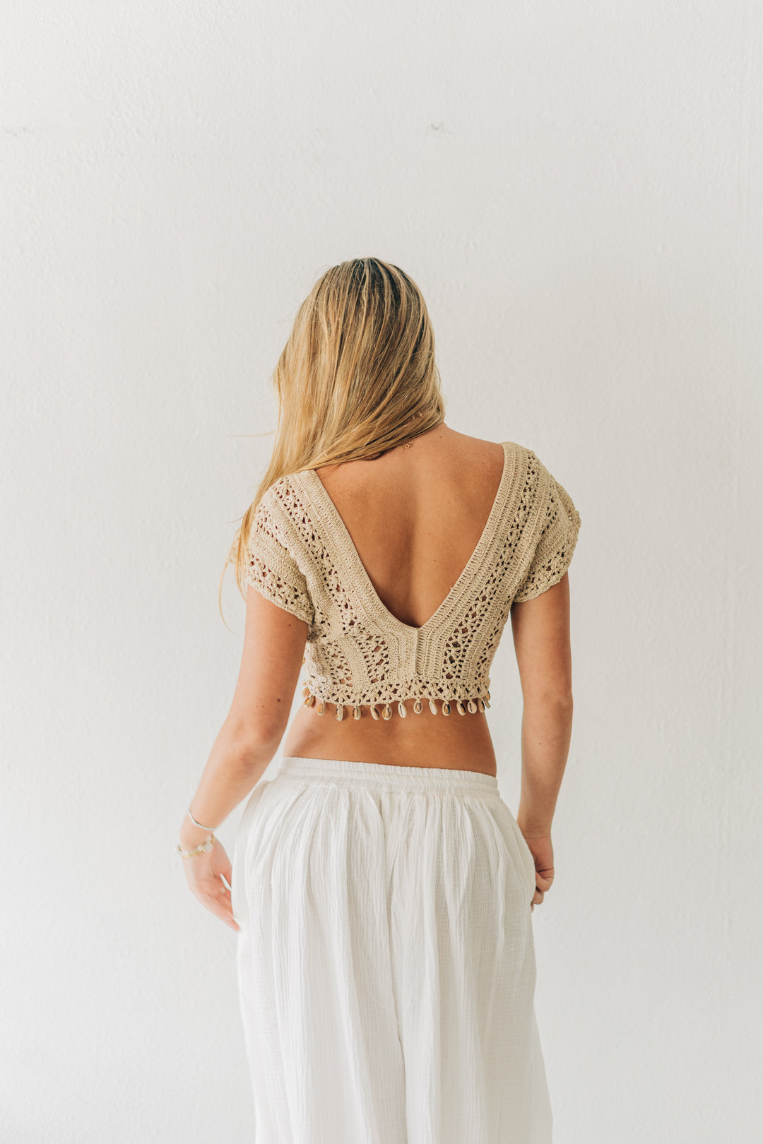 Woman wearing a beige lace top and white pants against a plain background