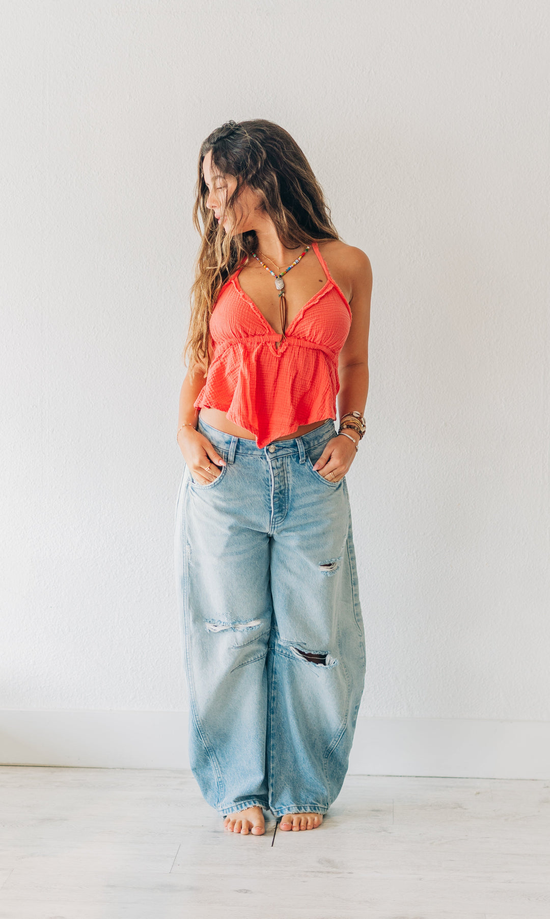 Woman wearing a coral top and light blue jeans standing in a room with white walls and minimal decor.