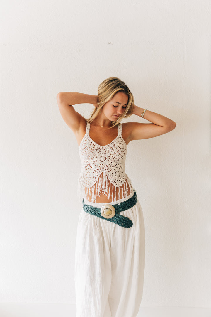 Woman wearing a white lace top and long white pants with a green belt against a plain background
