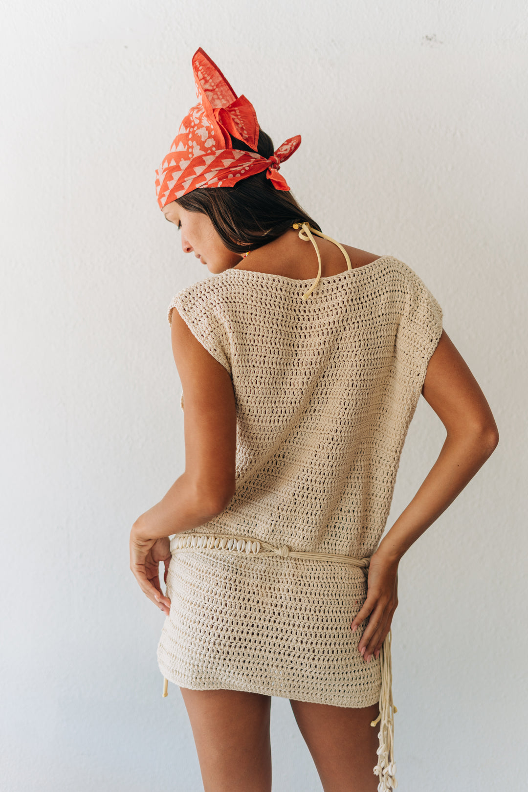 Woman wearing a beige crochet dress and red patterned headscarf against a white background