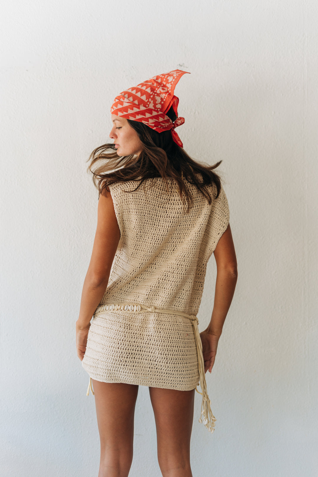 Woman wearing a beige crochet dress and red patterned headscarf against a white background