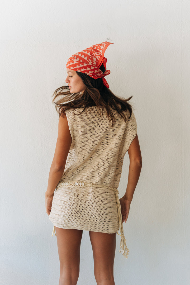 Woman wearing a beige crochet dress and red patterned headscarf against a white background