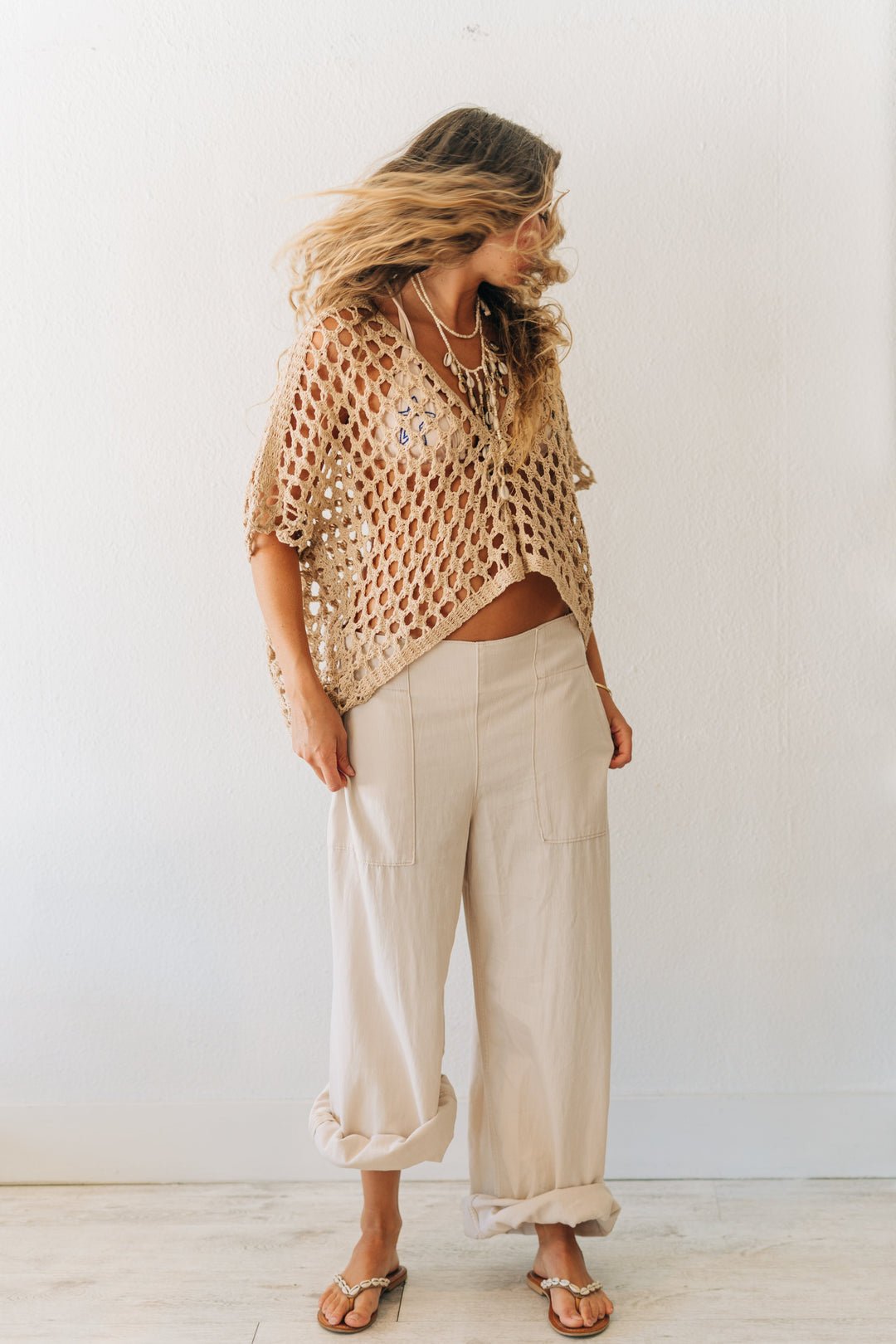 Woman wearing a beige knitted top and light-colored pants against a white wall.