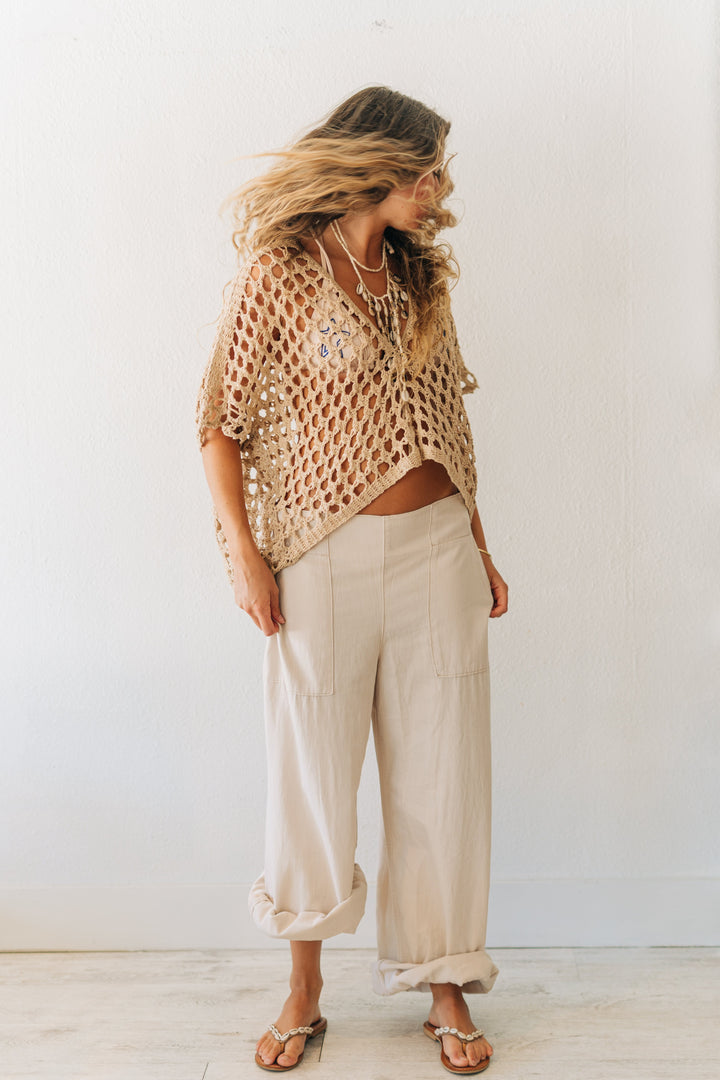 Woman wearing a beige knitted top and light-colored pants against a white wall.