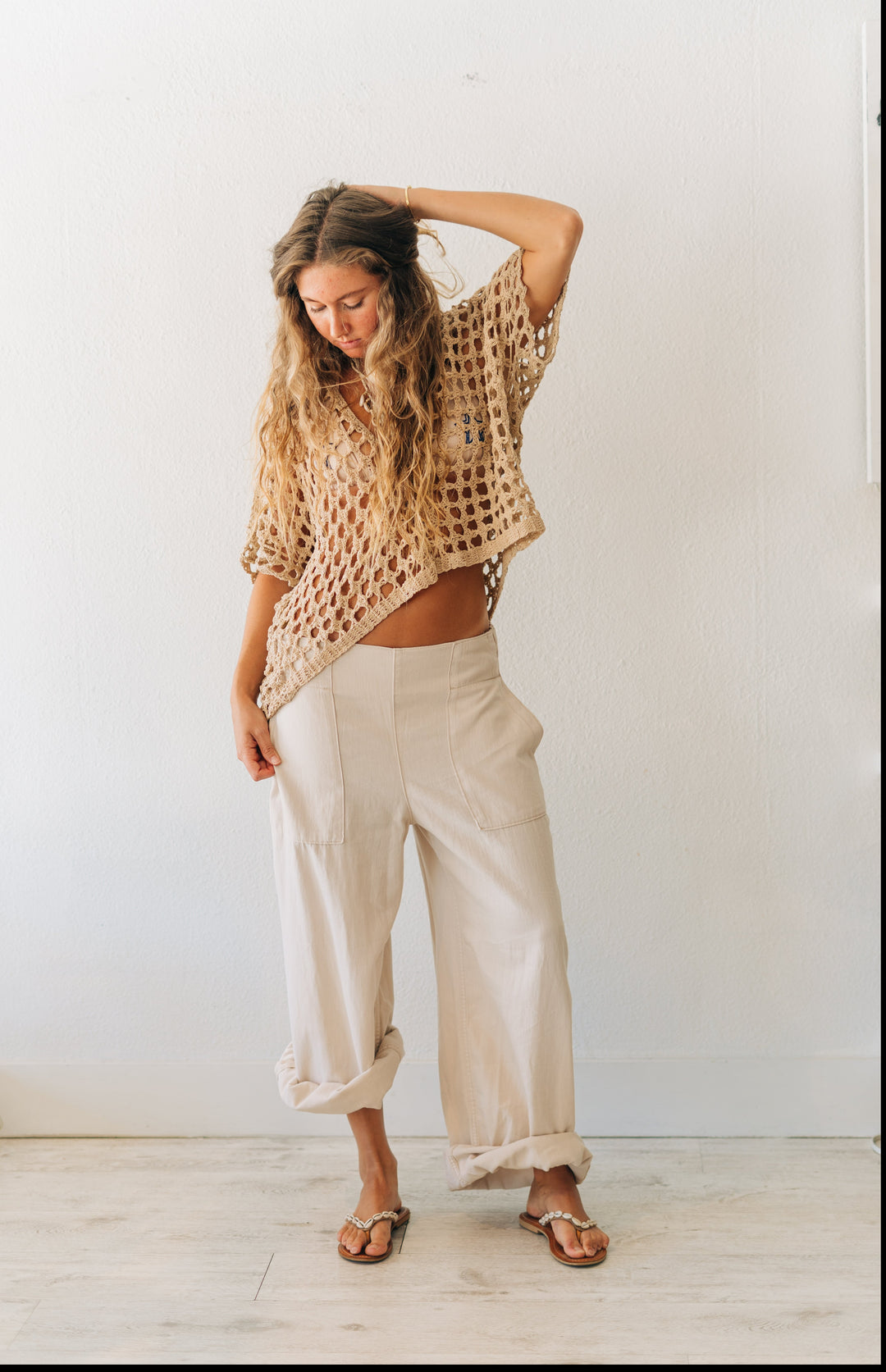 Woman wearing a beige crochet top and wide-leg pants against a white wall.