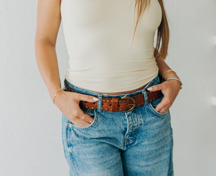 Person wearing a white top and blue jeans with a brown belt, standing against a neutral background.