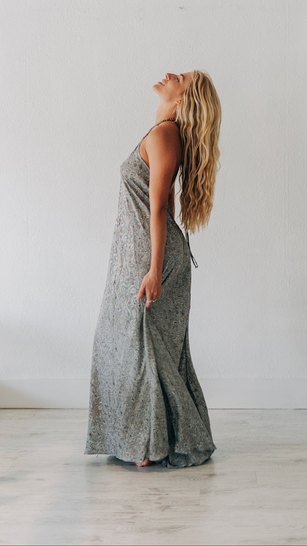Woman in a long gray dress standing in a room with a white wall and wooden floor.
