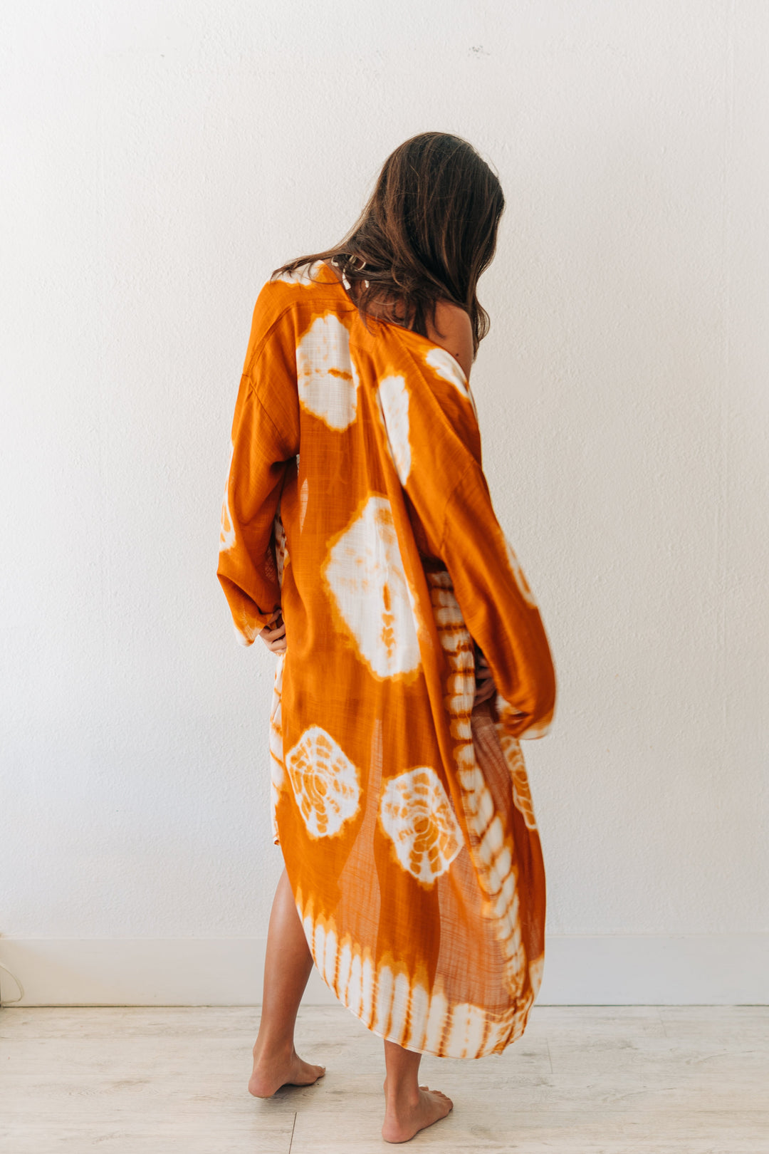 Woman wearing a long, orange tie-dye dress against a white wall.