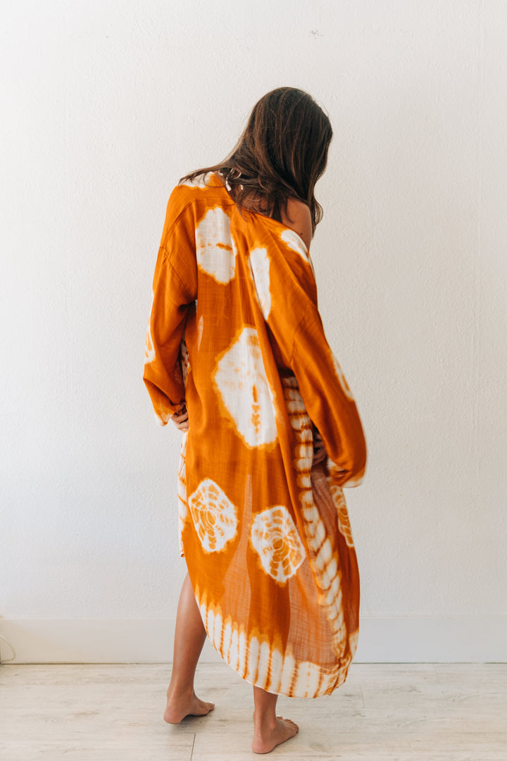 Woman wearing a long, orange tie-dye dress against a white wall.