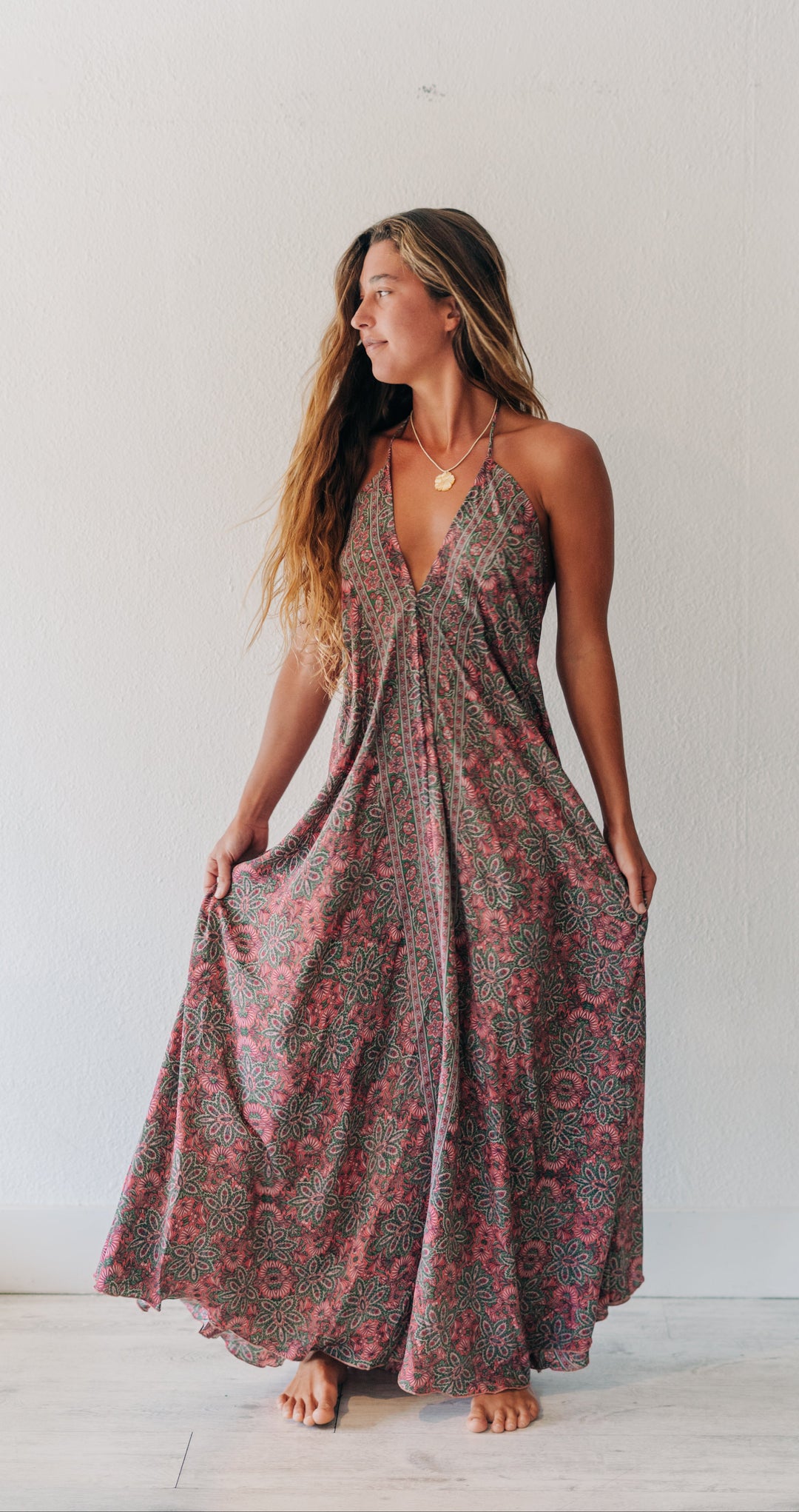 A woman wearing a sleeveless, floor-length bohemian maxi dress with a red and green floral pattern on a dark background, standing barefoot against a white wall.