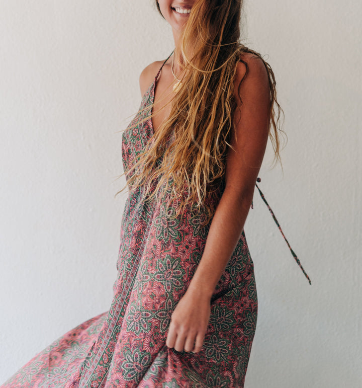 A woman with long, wet brown hair smiling while wearing a sleeveless, flowy maxi dress with a pink, green, and gray bohemian pattern, standing against a plain off-white wall.