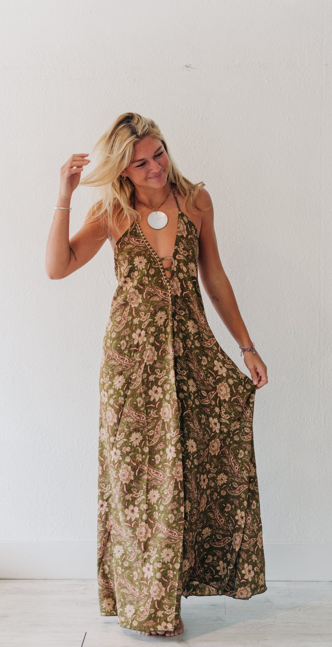 A woman modeling a sleeveless, floor-length floral maxi dress in olive green and beige tones, standing in a bright indoor setting with a white wall and light floor.
