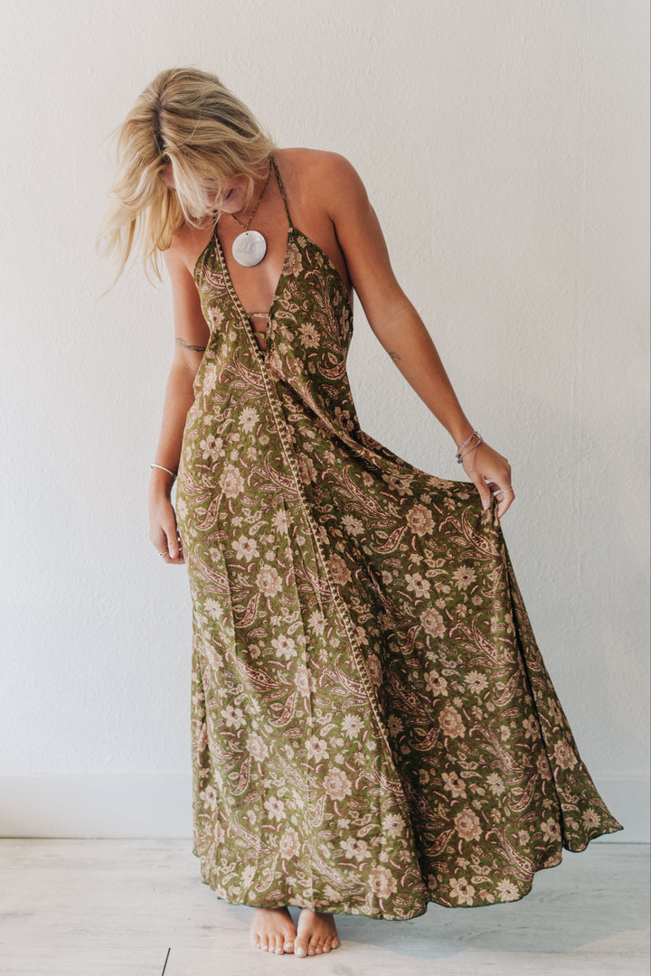 A woman wearing a sleeveless olive green maxi dress with a pink and beige floral pattern, standing barefoot against a white wall and holding out the side of the dress to show its flow.