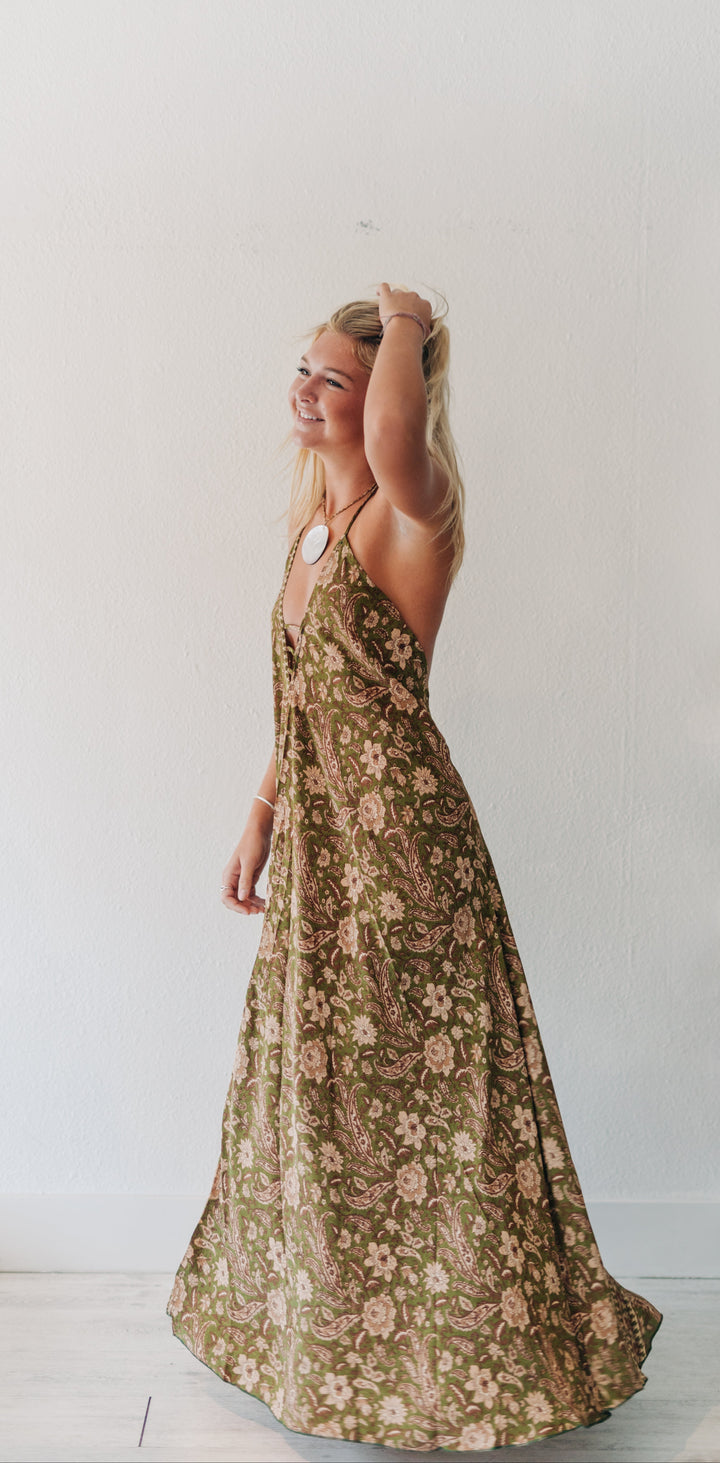 A woman wearing a sleeveless, floor-length maxi dress with a brown and beige floral pattern, standing in profile against a white wall.