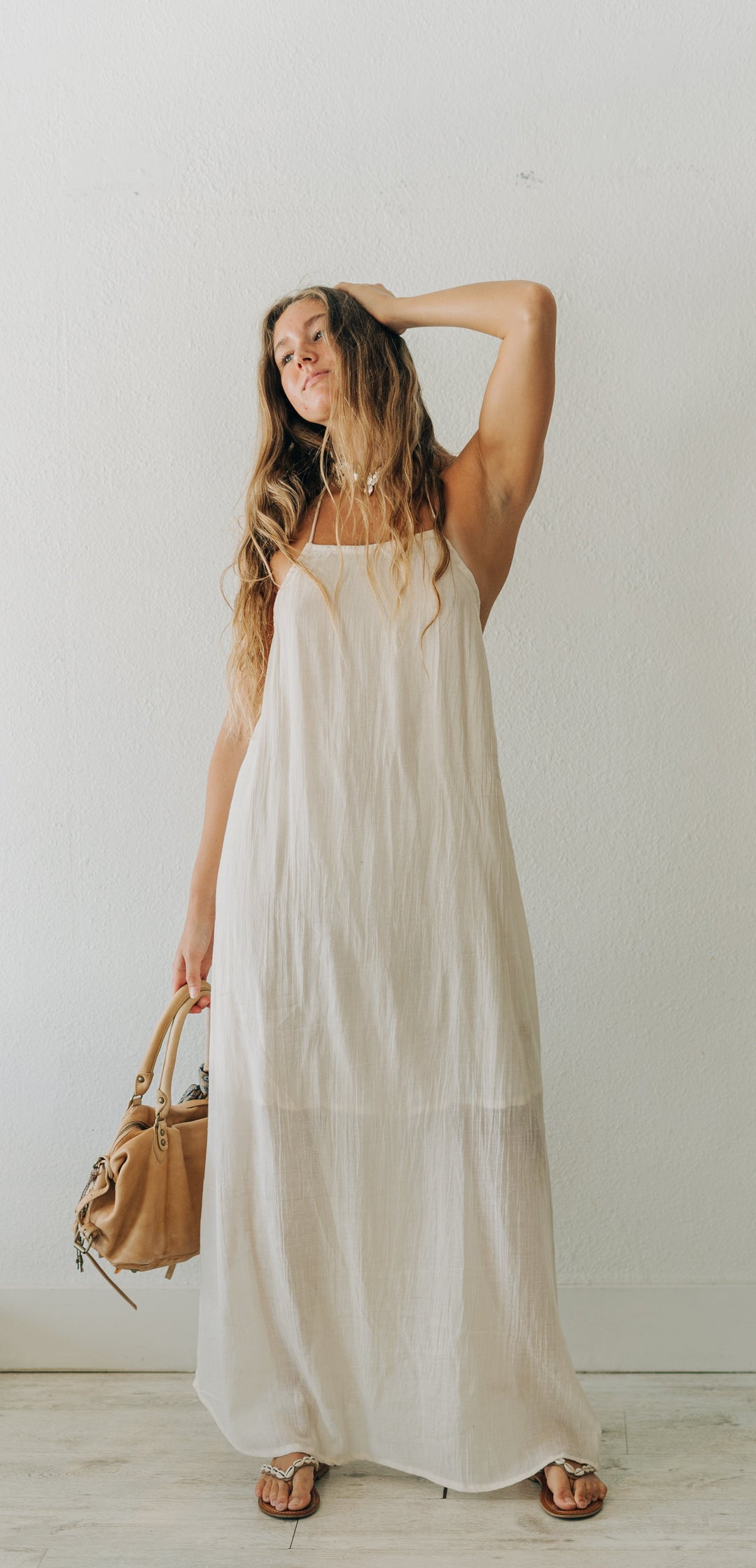Woman in a white dress holding a brown handbag against a plain white wall.