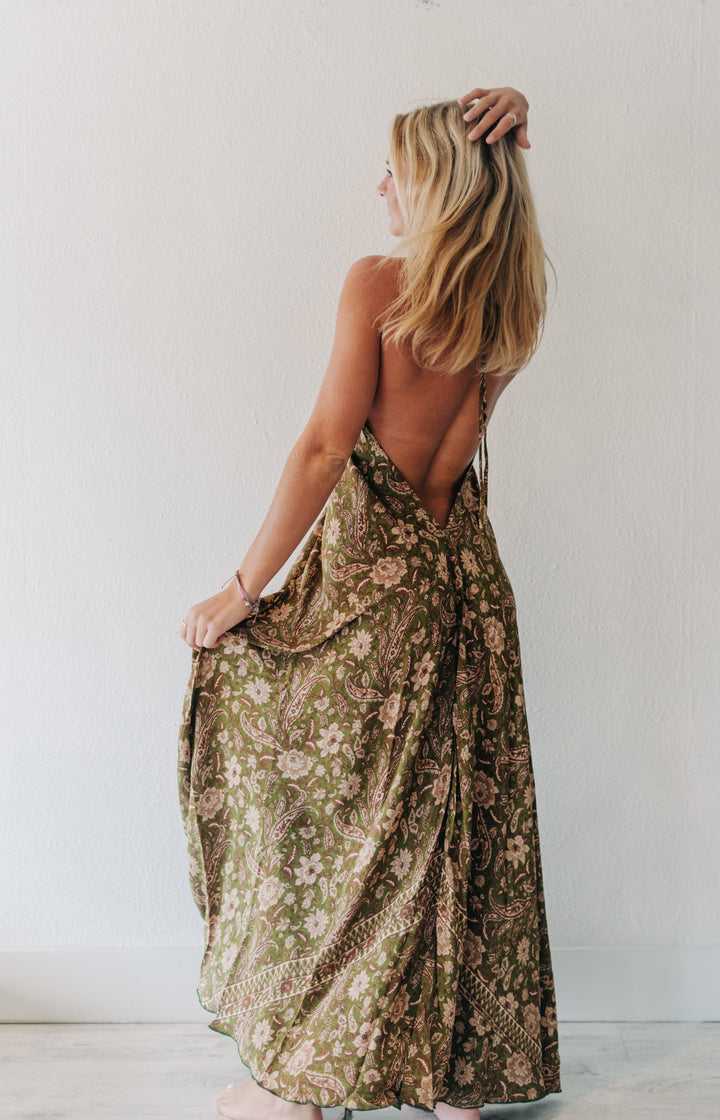 A woman with blonde hair is shown from behind wearing a long, open-back maxi dress with a brown and beige floral pattern, standing against a plain white wall.