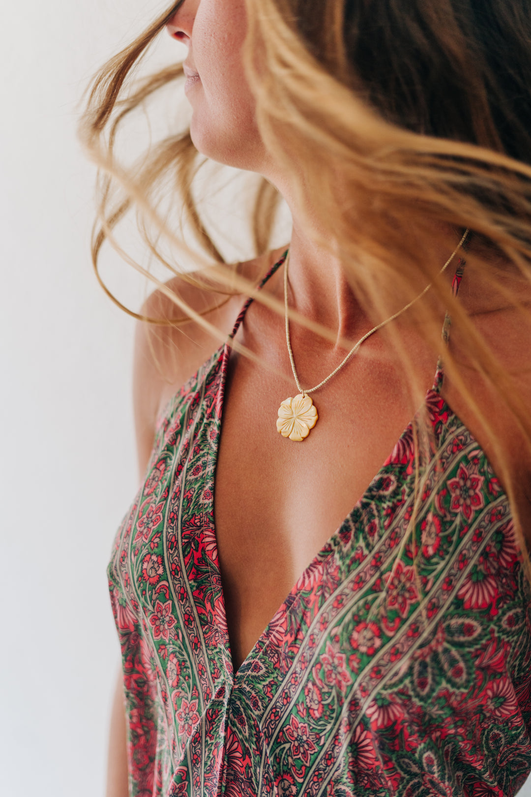 A close-up lifestyle shot of a woman wearing a delicate gold flower pendant necklace and a red and green patterned halter top against a bright white background.