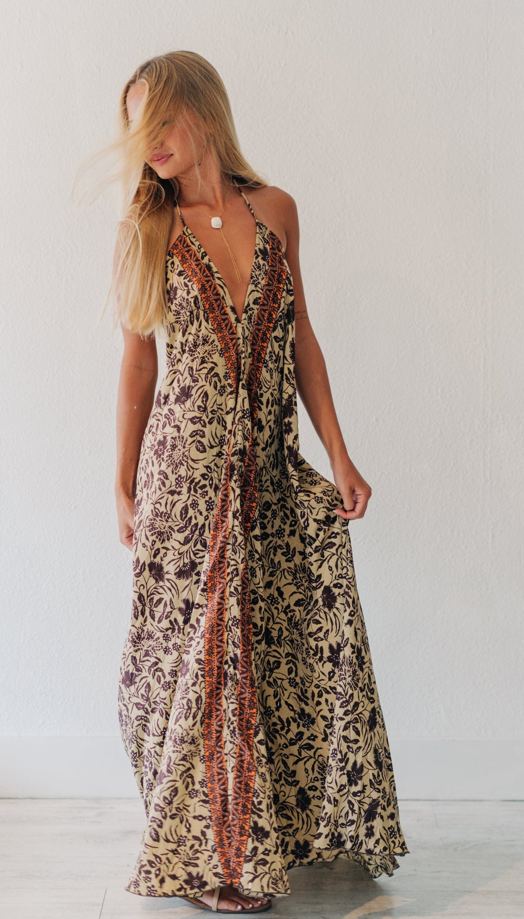 A woman modeling a long halter-neck maxi dress with a brown and beige floral pattern and a rust-orange vertical stripe detail down the front, set against a plain white wall.