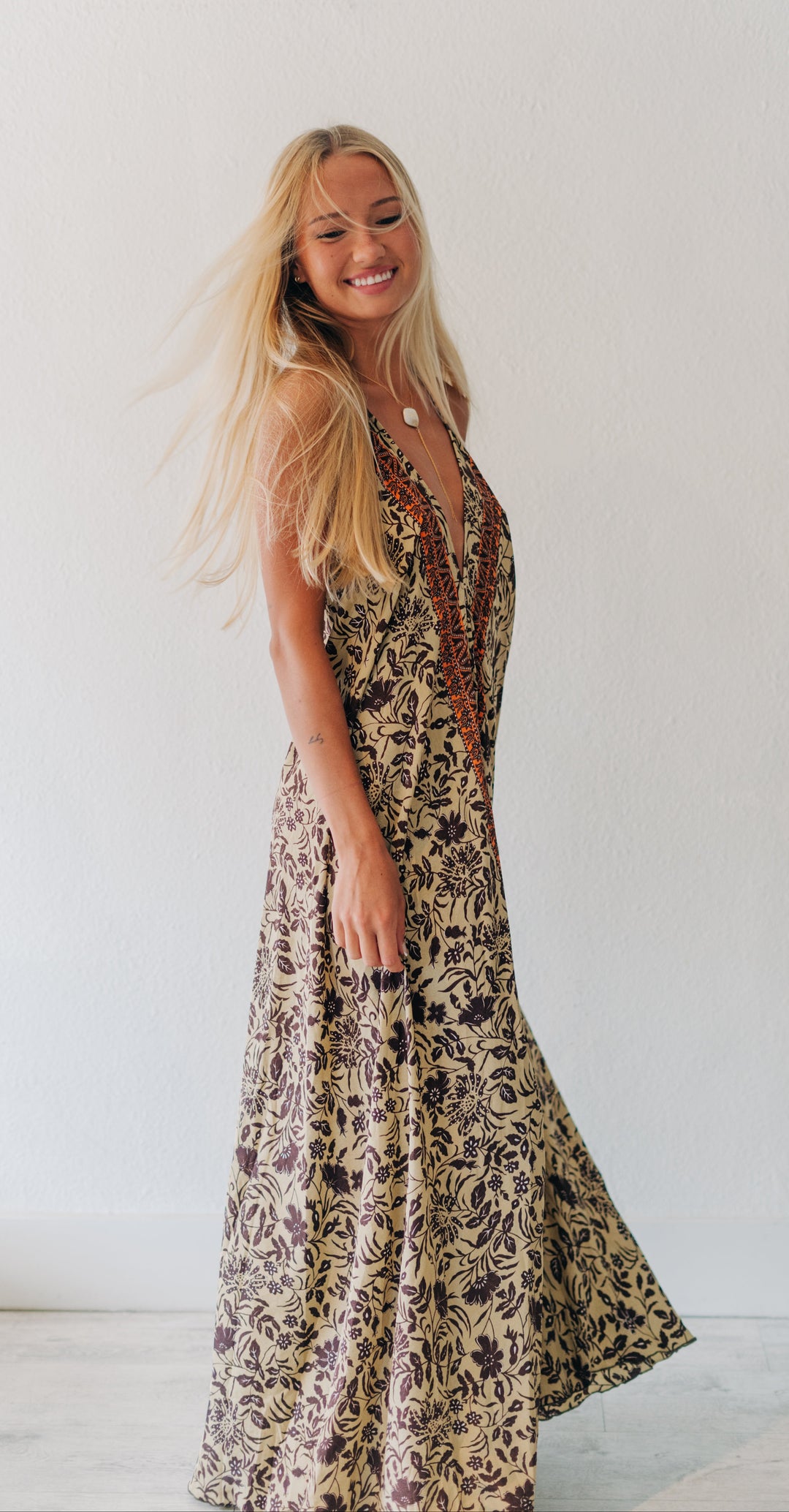 A woman with long blonde hair wearing a sleeveless bohemian-style maxi dress with a brown and beige floral pattern, standing in a bright white room.