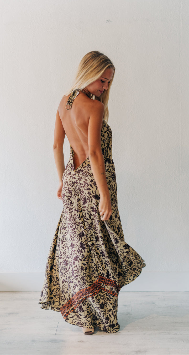 A woman wearing a sleeveless, open-back maxi dress with a brown and beige floral pattern, photographed from behind in a bright, minimalist room.