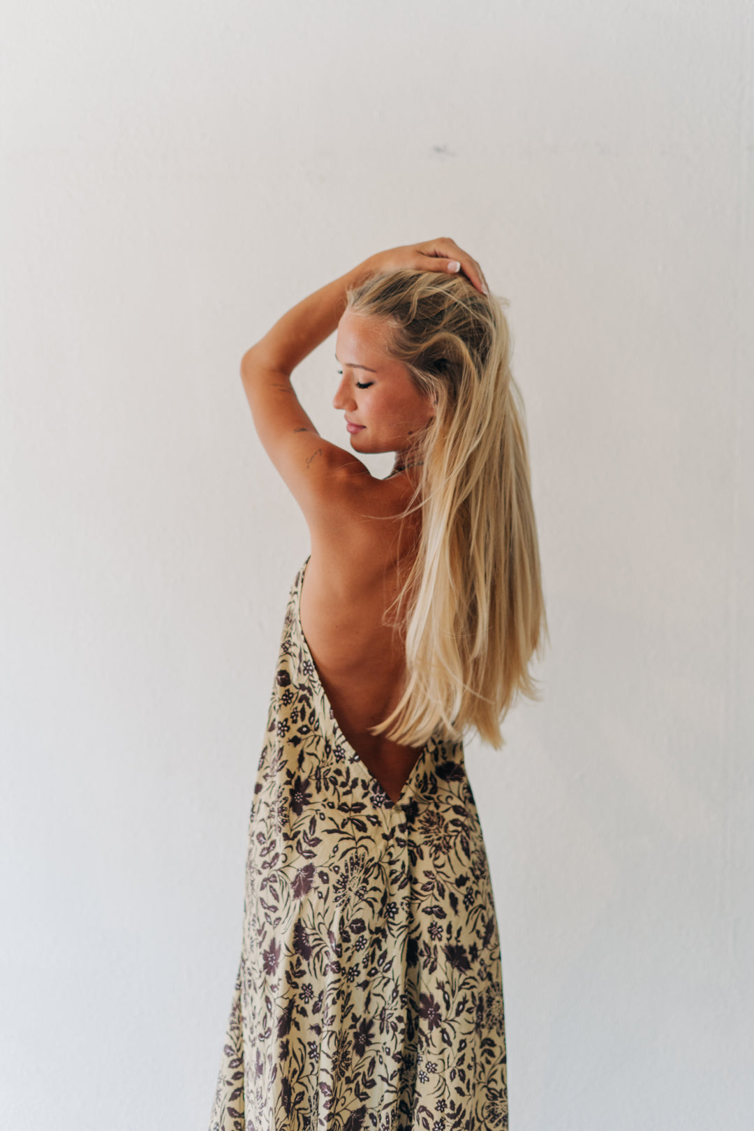 A woman with long blonde hair is shown from the back wearing a sleeveless, open-back dress with a beige and brown floral pattern, posing against a plain white wall.
