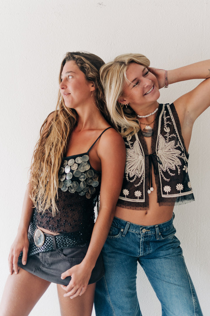 Two women posing back-to-back in bohemian-style clothing against a white wall. The woman on the left wears a black lace camisole with silver coin embellishments and a studded belt, paired with dark denim shorts.