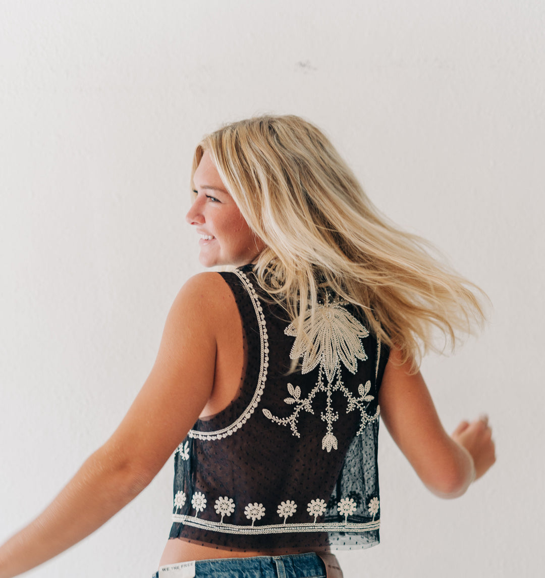 A woman with blonde hair is shown from behind wearing a black sleeveless crop top with white floral embroidery and blue jeans, set against a plain white wall.