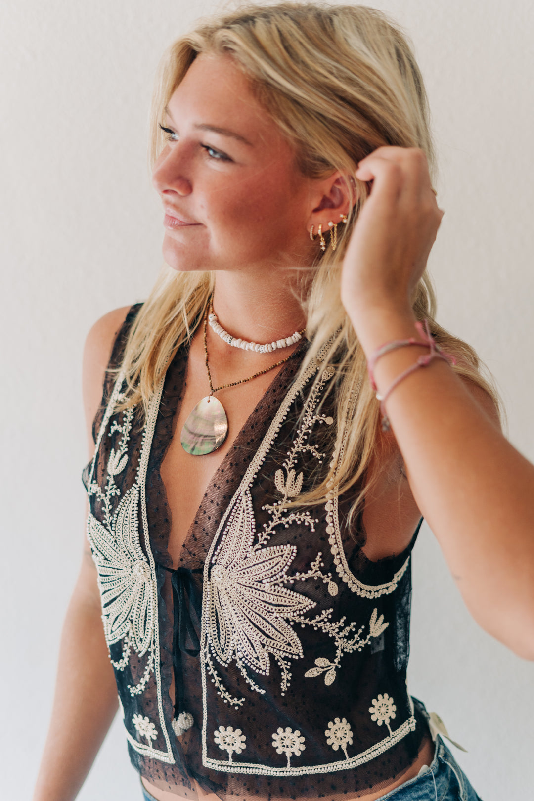 A blonde woman wearing a sleeveless black mesh vest with intricate white floral embroidery, layered with a pearl choker and a large green pendant necklace.