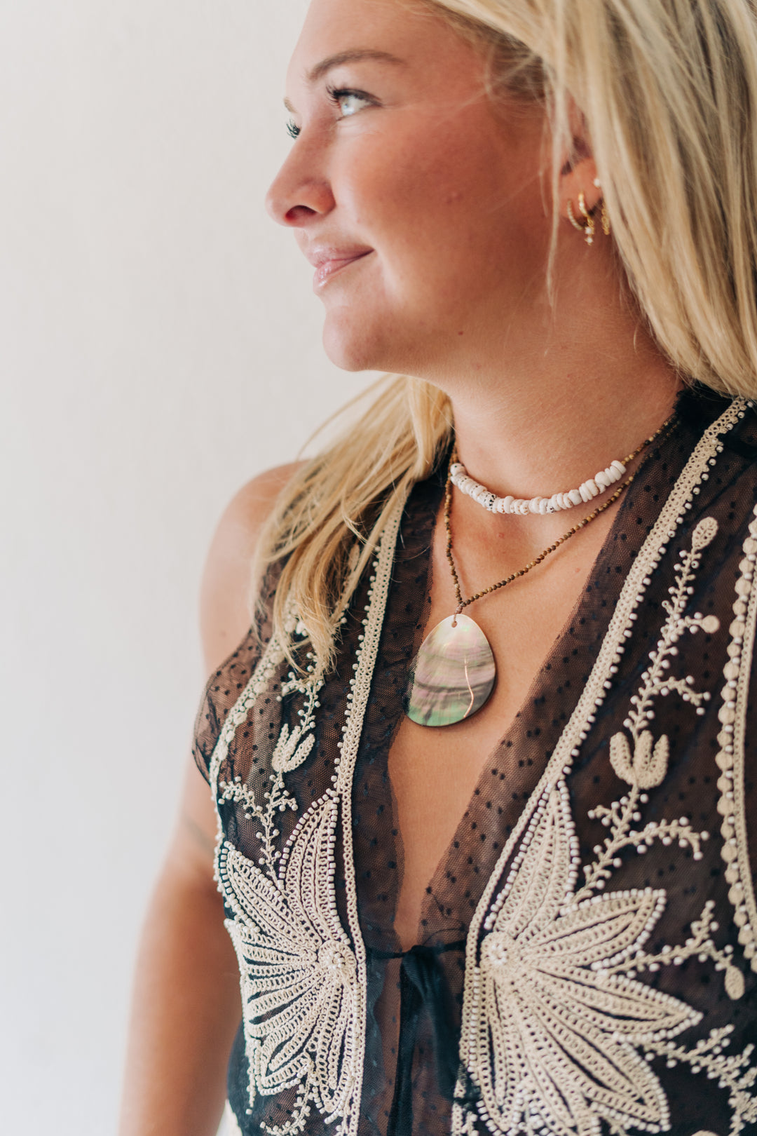 A close-up side profile of a woman wearing layered necklaces, including a prominent teardrop-shaped abalone shell pendant on a chain, a pearl choker, and a delicate gold hoop earring, paired with a sheer black embroidered top.